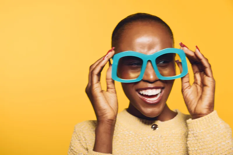 Smiling African woman with dark skin wearing oversized blue glasses and a beige sweater poses against yellow background. Concept of fashion, joy, and vibrant lifestyle.