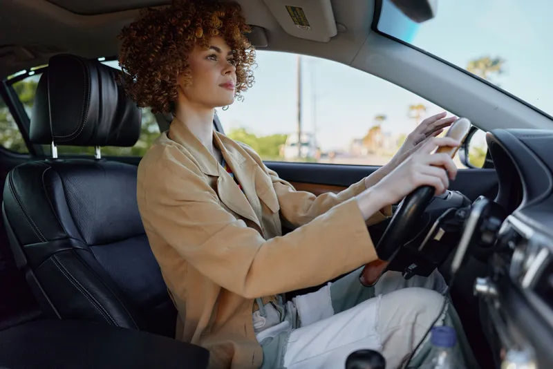 Candid woman driving a car, focused expression, sunlit interior, retro film color vibe, casual blazer, natural light, minimal background, lifestyle moment