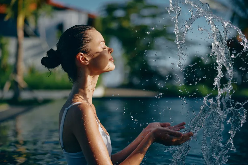Elegant woman stands beside a sunlit fountain, feeling calm as water sprays her face, enjoying outdoor relaxation and refreshing moment by a tranquil pool, capturing a serene lifestyle mood