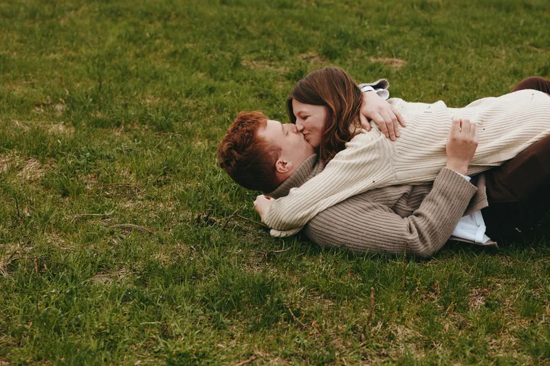A couple in sweaters share a kiss on green grass, capturing a candid, intimate outdoor moment. A warm lifestyle scene depicting love, togetherness, and calm in a natural setting.