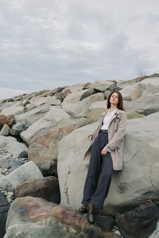 Young woman in casual outfit stands on large rocks near the shore under cloudy sky. Relaxed pose and natural setting create mood of calm and contemplation outdoors.