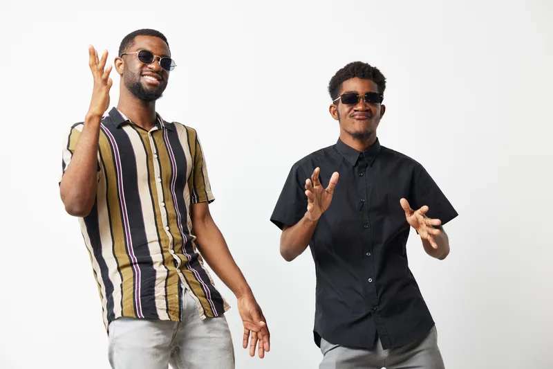 Two stylish black men in trendy sunglasses showcasing joyful dance moves. Their vibrant outfits enhance the lively mood, celebrating togetherness and friendship.