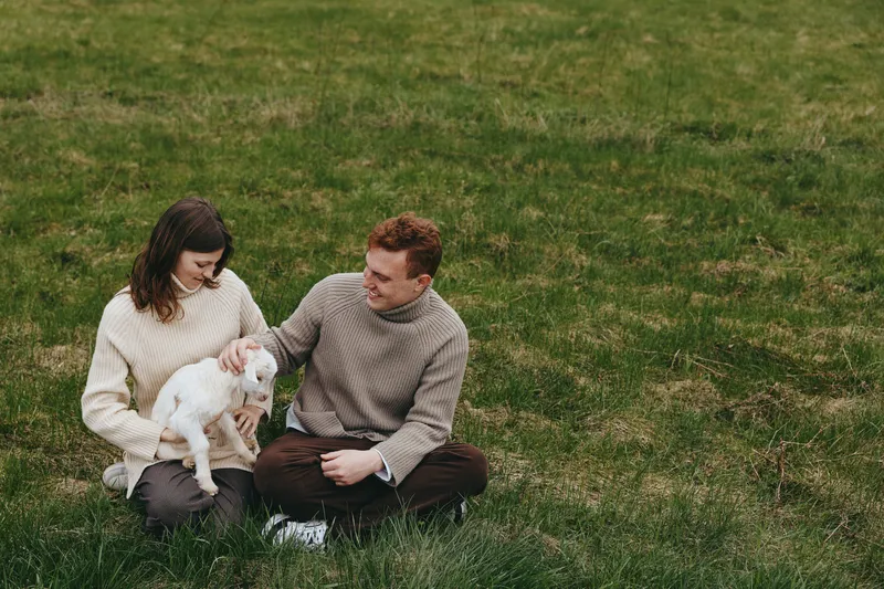 A couple with a goat kid sits on grass in a sunlit field, sharing a tender moment. A lifestyle scene highlighting family bonds, companionship with animals, and serenity outdoors.