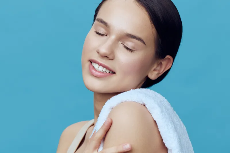 A young woman with glowing skin, wrapped in a towel, smiles gracefully against a serene blue background, conveying beauty and confidence in self care and skincare routines
