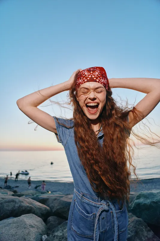 Lifestyle woman in modern boho-western denim street style laughing joyfully with wind-blown hair at the beach during golden hour in vibrant film color tones.