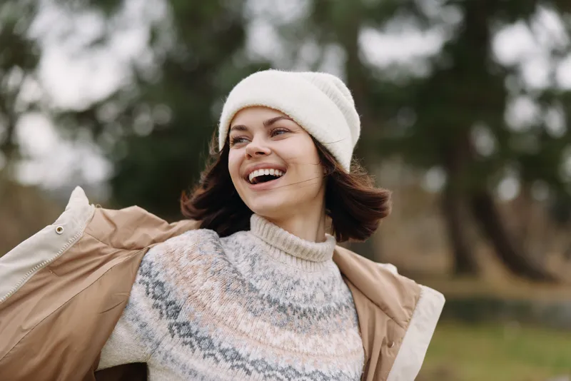 Joyful woman outdoors wearing a fashion sweater and beanie, smiling and carefree in a windy park scene with autumn light