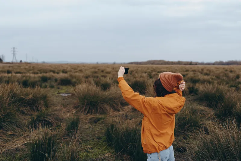 Woman lifestyle outdoors in a grass field, orange jacket and knit hat, cloudy sky behind windy air, she raises one arm posing for a photo, distant power lines, freedom mood today