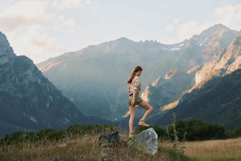 Woman hiking on a rock in a grassy meadow with towering mountains in the background, capturing an outdoor lifestyle moment and a sense of exploration and adventure.