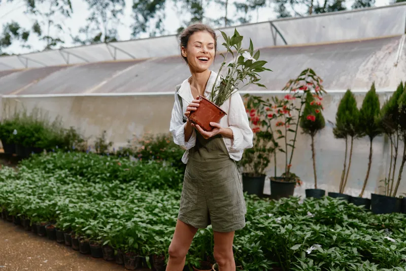 Young woman holding a potted plant in a greenhouse, radiating joy and connection with nature, wearing casual outfit suitable for gardening, captured in natural light