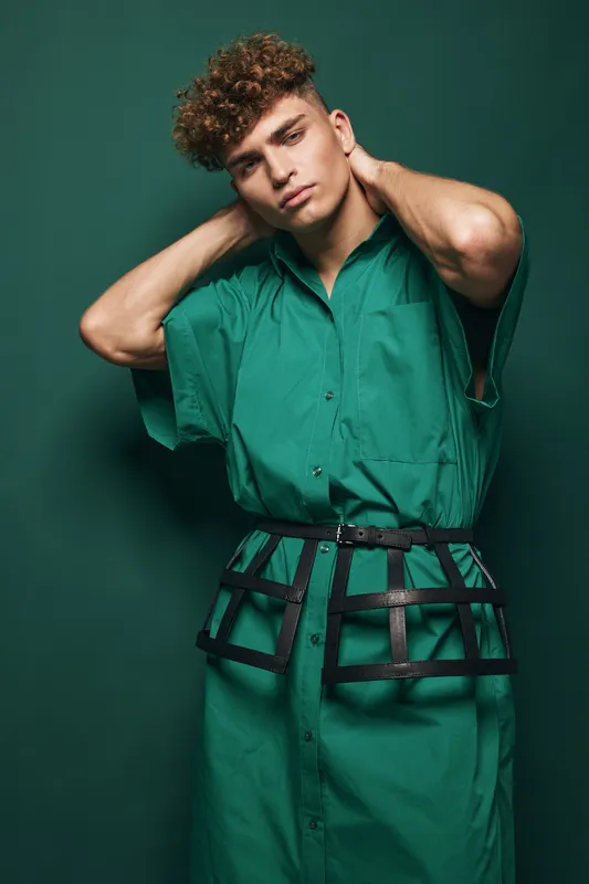 Young man with curly hair wearing green oversized shirt and cage belt posing on dark green background for fashion, hairstyle, style, casual, urban and portrait concept.