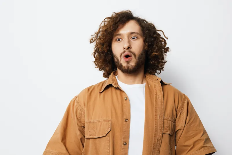 Young man with curly hair expressing surprise in casual clothing against a neutral background, ideal for capturing emotions and everyday moments