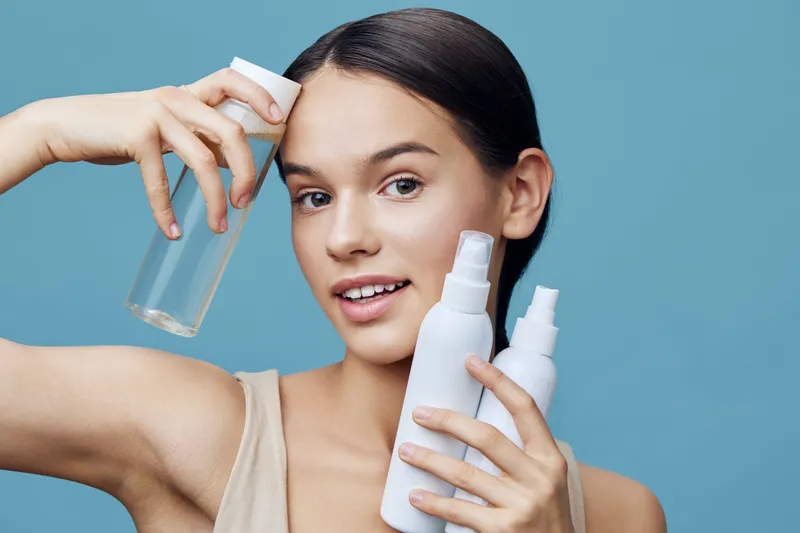 Young woman holding skincare products, showcasing healthy skin and beauty routine, on a light blue background with natural expression and soft lighting