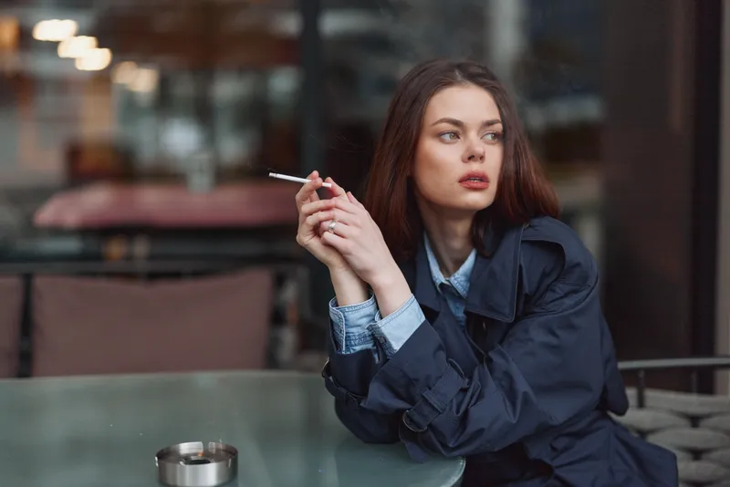 Young woman with long brown hair wearing a dark coat smokes a cigarette while sitting at a glass table in a dimly lit indoor cafe.