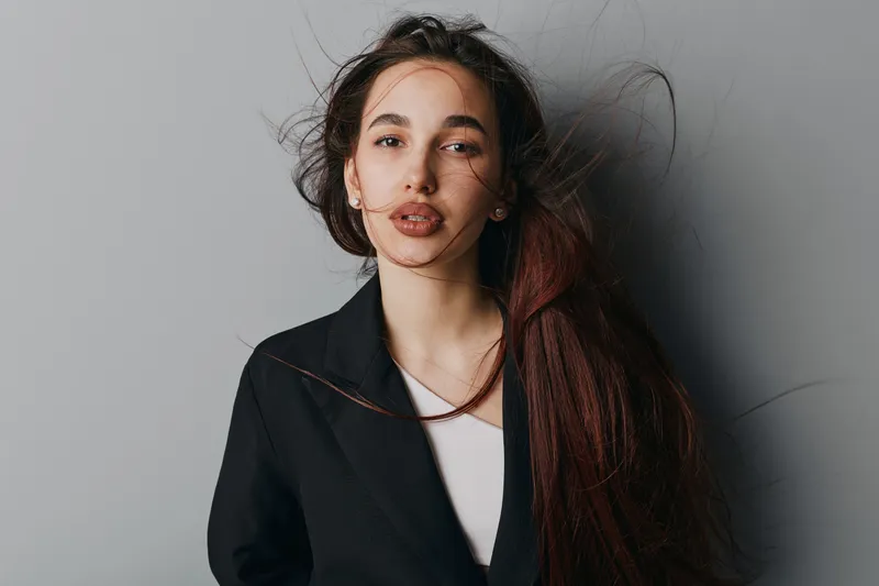 Young woman with long hair and natural makeup posing in black jacket against gray background. Female model with serious expression and loose hair in casual fashion portrait.