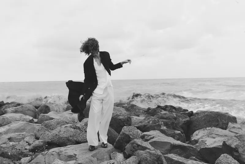 Woman walking on rocks along the shoreline, windblown hair, jacket open and white pants, sea horizon in the background, bold coastal moment in monochrome