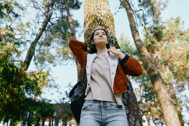 A woman stands outdoors beside a tall tree in a park, looking upward with a calm smile, wearing a sweater and jacket, basking in daylight among autumn leaves and gentle breeze.