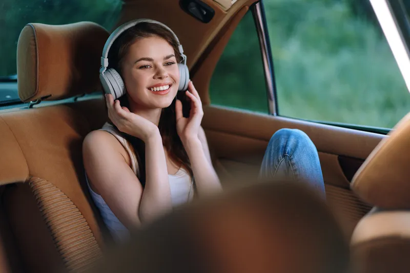 Young woman enjoying music in car with headphones, smiling happily in a casual outfit, surrounded by natural green background, conveying a joyful and relaxed mood.