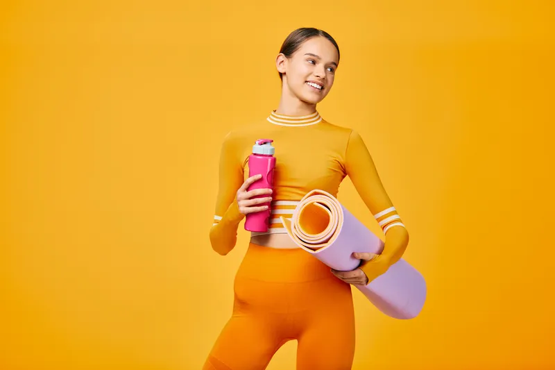 Woman fitness enthusiast stands against a vibrant orange backdrop, holding a pink water bottle and a rolled yoga mat, ready for workout and sport training in a dynamic athletic pose.