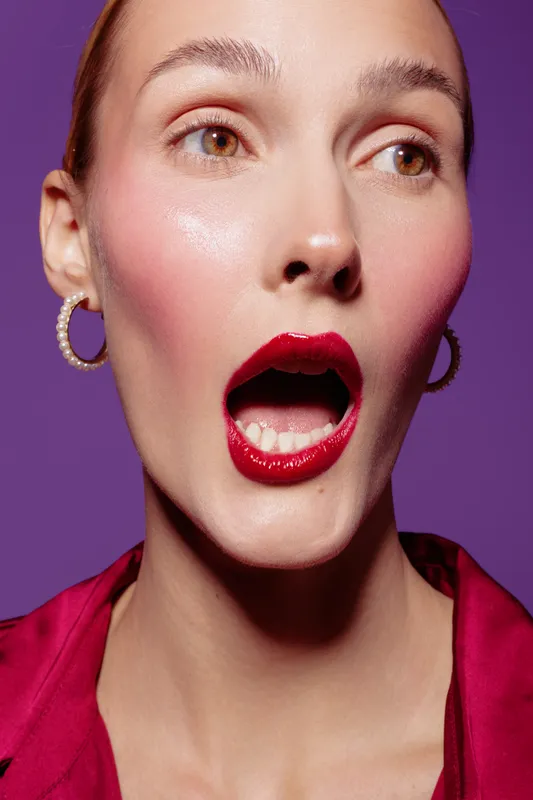 Close-up of surprised woman with red lipstick and hoop earrings wearing a red blouse on purple background. Concept of beauty, fashion, makeup, and emotional expression.