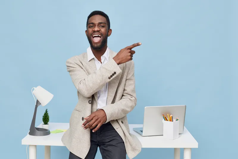 Vibrant smiling man in business casual outfit pointing to the side, sitting at modern office desk with laptop, lamp, and potted plant against blue background, cheerful professional workplace concept.