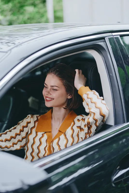 A cheerful woman sits in a car, smiling softly with one hand through her hair, enjoying a relaxed moment behind the wheel.