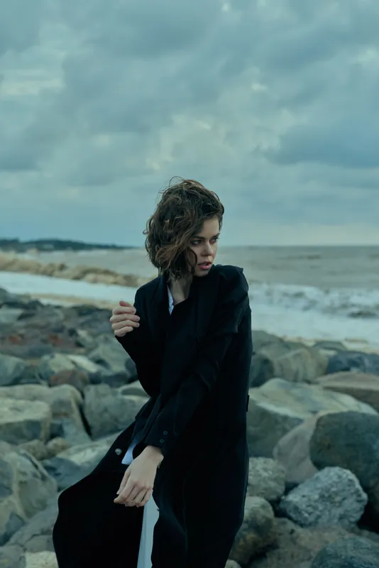 A moody fashion shot of a model in a dark coat on a rocky beach under stormy skies, capturing wind, texture, and movement along a coastal seascape for an editorial, atmospheric outdoor tale