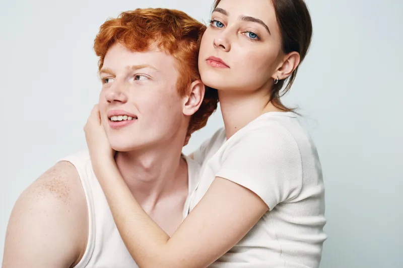 Couple with woman hugging man wearing white tank tops looking away closeup studio, intimacy and connection between young adults in casual outfits on light background.