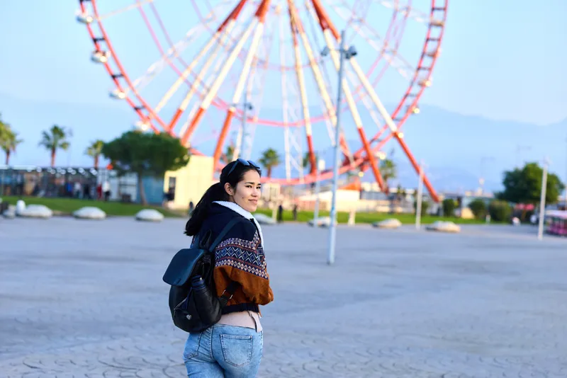 Woman with backpack at an amusement park near a large ferris wheel, casual sweater and jeans, turning toward the camera in an outdoor daytime scene