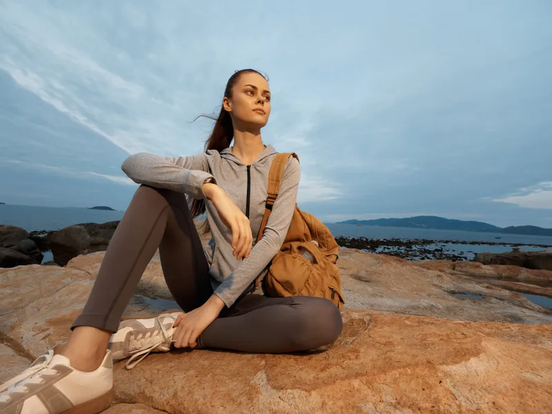 Young woman in sportswear sitting on rocks by the sea, promoting healthy lifestyle and outdoor adventures in serene blue tones