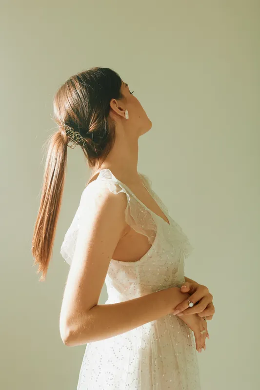 Young woman in a white lace dress poses with eyes closed and head tilted back. Natural light highlights her beauty and fashion style in a soft indoor setting.