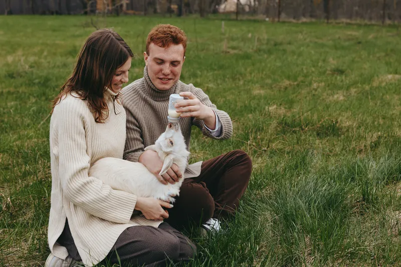 Candid couple outdoors in a grassy field, smiling while they cradle a small goat kid, cozy knitwear, sunlit moment, gentle breeze, natural colors, relaxed warmth, countryside backdrop