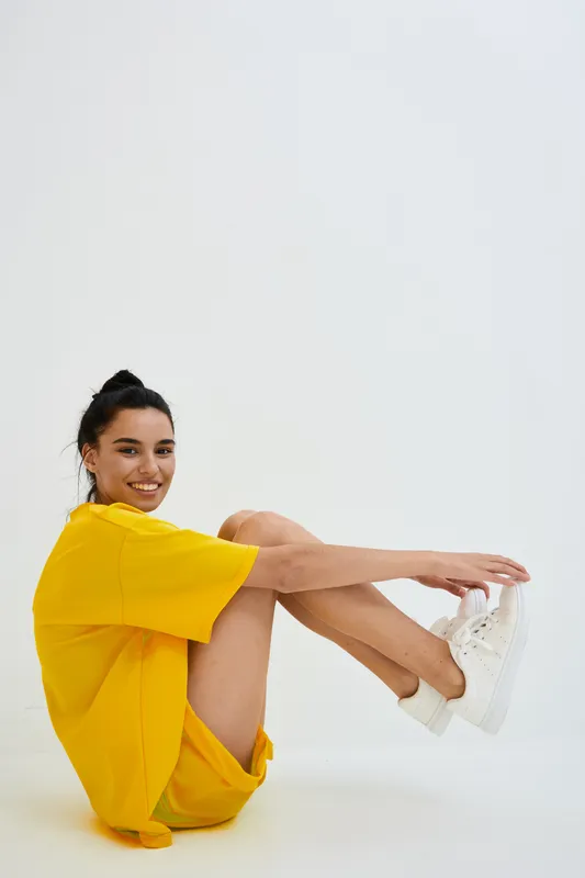 Smiling woman in yellow oversized T-shirt and white sneakers sitting on floor against white background with relaxed pose, casual style, and cheerful expression.