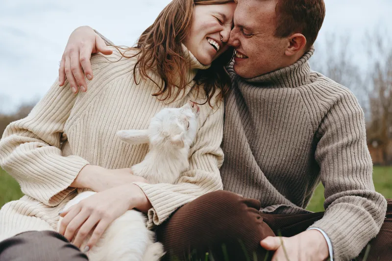Loving couple outdoors with a playful goat kid, sharing a warm moment on a grassy field. Warm natural light highlights happiness, bonding, and gentle affection in a casual lifestyle scene.