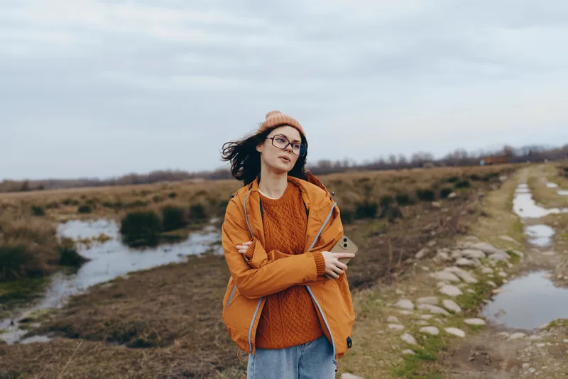 Woman lifestyle outdoors in countryside, wearing jacket and hat, standing on a rural walkway, arms crossed, cloudy sky, relaxed calm mood, natural colors, distant fields