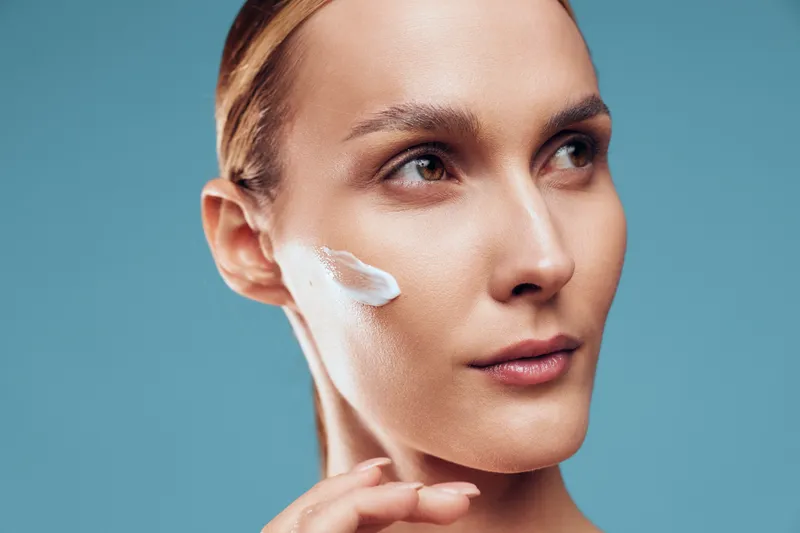 Closeup of young woman applying cream on clean face with smooth skin natural makeup and focused expression isolated on blue background beauty skincare concept.