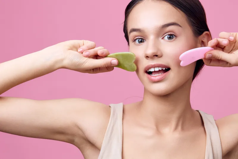 Young woman holding skincare tools on a pink background, showcasing a joyful expression, promoting facial massage and self care for healthy skin