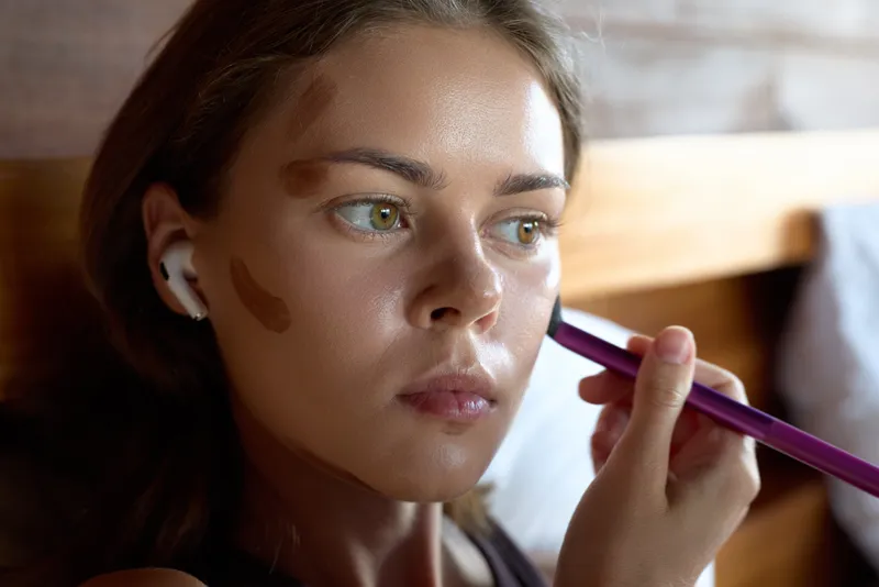 A young woman with vitiligo applies makeup with a brush while wearing wireless earbuds in a cozy indoor setting. Concept for beauty, self-care, and skin diversity.