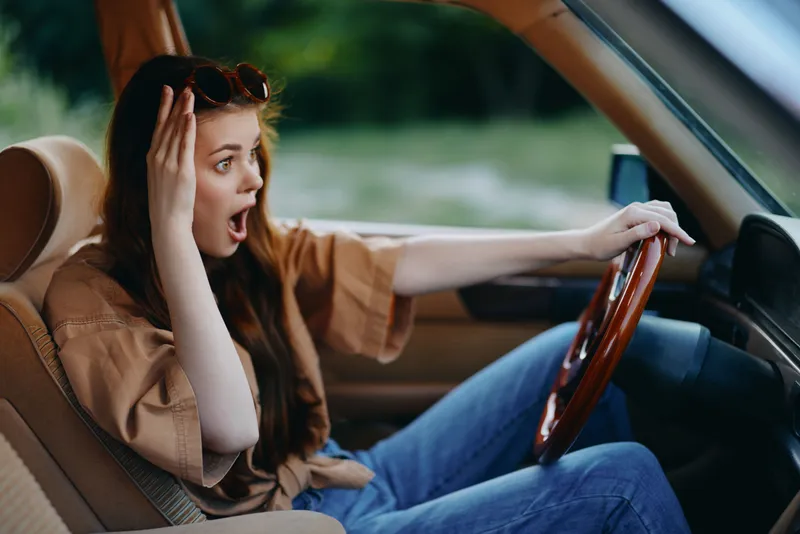 Shocked young woman driving in a car, expressing surprise with a worried look, wearing casual clothing and sunglasses, set in a serene outdoor background.