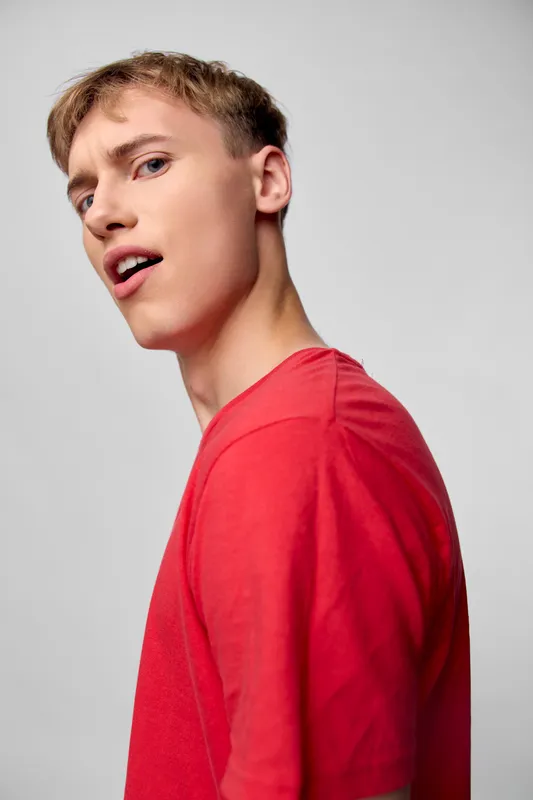 Man in a red shirt strikes a confident profile pose in a clean studio, showcasing casual fashion and modern style, bright lighting highlighting features and a relaxed, expressive mood.