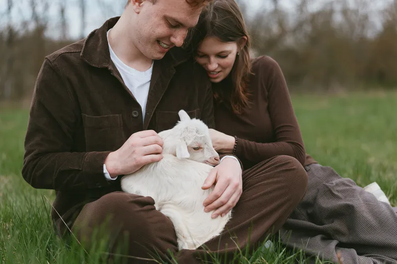 Couple sits on grass in a meadow with a gentle goat kid resting on their laps, warm affectionate moment outdoors, casual rustic clothing, calm atmosphere, natural light, true lifestyle scene with