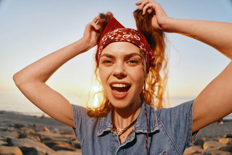 Lifestyle woman in modern boho-western denim street style smiling outdoors with red bandana and warm film color during sunset by rocky shore.