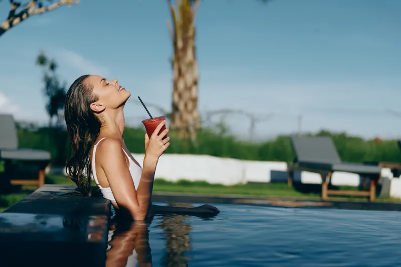 Young woman with curly hair enjoys a refreshing drink while relaxing in an outdoor pool under clear blue sky, embracing a peaceful lifestyle moment.