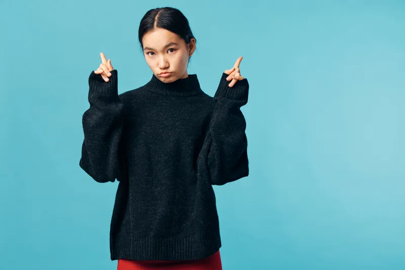 Young woman wearing oversized black sweater on blue background posing with pout and crossed fingers for luck or hope, casual style and confident expression in studio portrait.