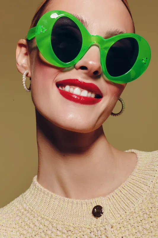 Close-up of smiling woman wearing large green sunglasses and red lipstick. She has hoop earrings and a beige knitted sweater. Concept for fashion, style, and summer accessories.