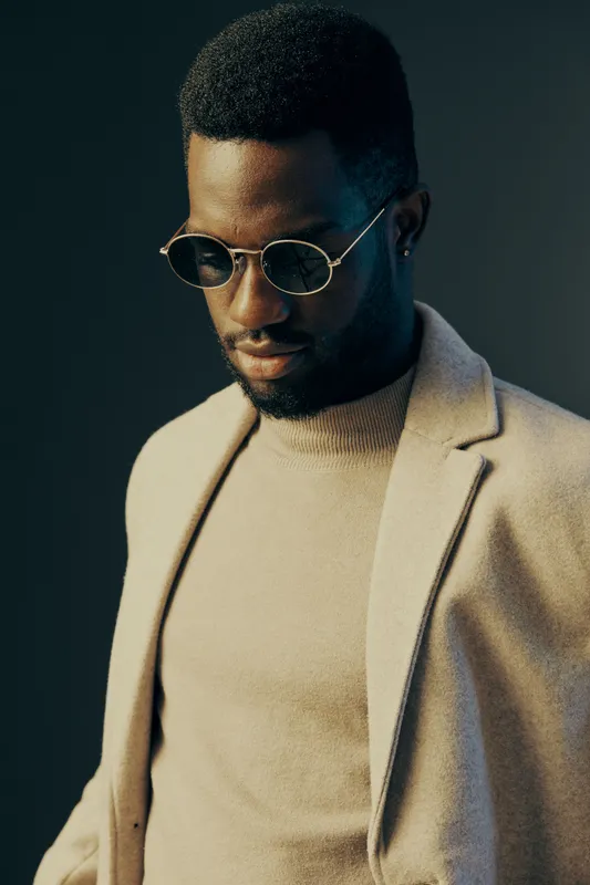 Stylish African American man in sunglasses and a beige coat against a dark background, showcasing modern fashion trends and confidence