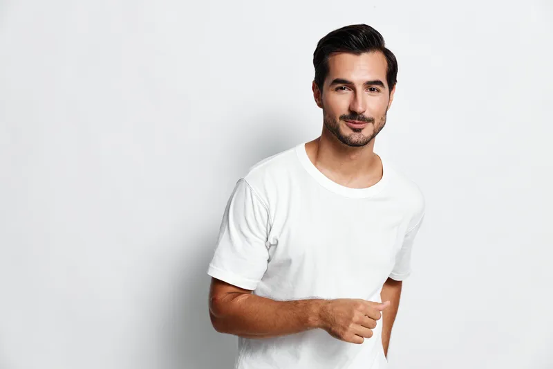 Smiling young man wearing a white t-shirt stands confidently, looking directly at the camera with a friendly expression on his face against a plain background.