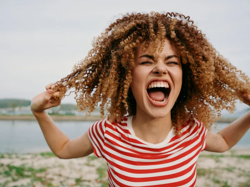 Close-up of a joyful young woman shouting with arms raised, curly hair bouncing, wearing a red and white striped shirt, outdoors near a sandy shore and calm water.