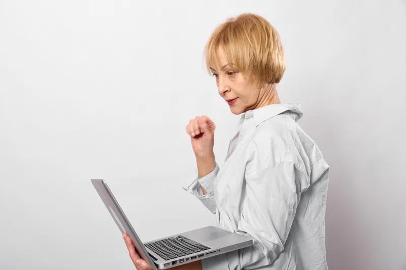 Senior woman in medical coat works with laptop, clenched fist showing anger, short blonde hair, isolated on white studio background, healthcare concept.