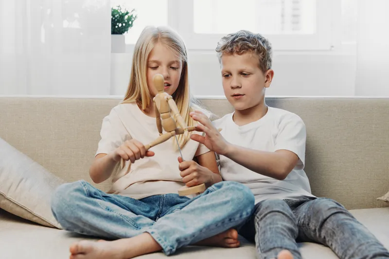 Two children sitting on a sofa playing with a wooden artist mannequin in a bright living room. Concept of childhood, creativity, and learning through play.