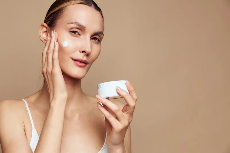 Young woman applies cream to her face, showcasing a simple skincare routine with a smooth glow, neutral background, soft lighting, clear skin, and a calm, focused expression.